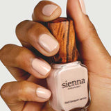 A hand with neatly manicured nails painted in pale nude holds a bottle of Sienna Byron Bay Nail Polish - Posy, featuring the brand's signature wooden cap, against a plain background.