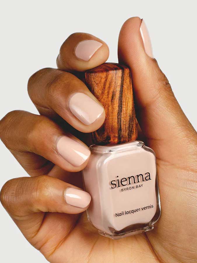 A hand with neatly manicured nails painted in pale nude holds a bottle of Sienna Byron Bay Nail Polish - Posy, featuring the brand's signature wooden cap, against a plain background.