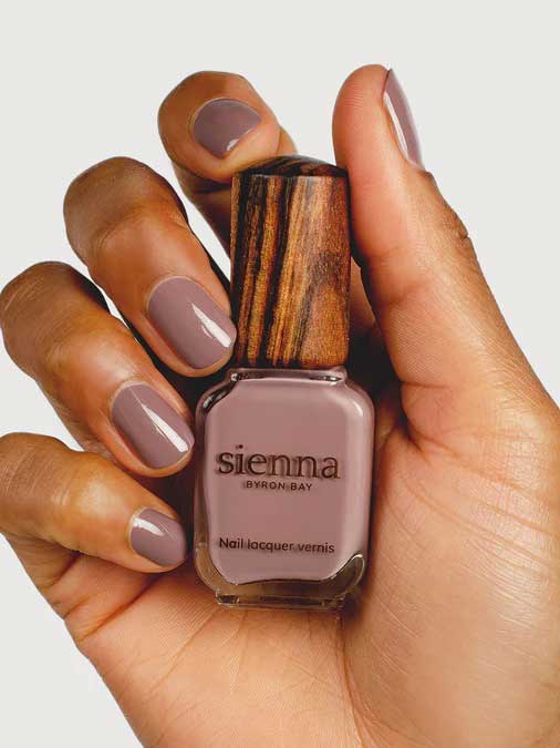 A hand with neatly manicured nails painted mauve holds a Sienna Byron Bay Nurture nail polish bottle, featuring its distinctive wooden cap.