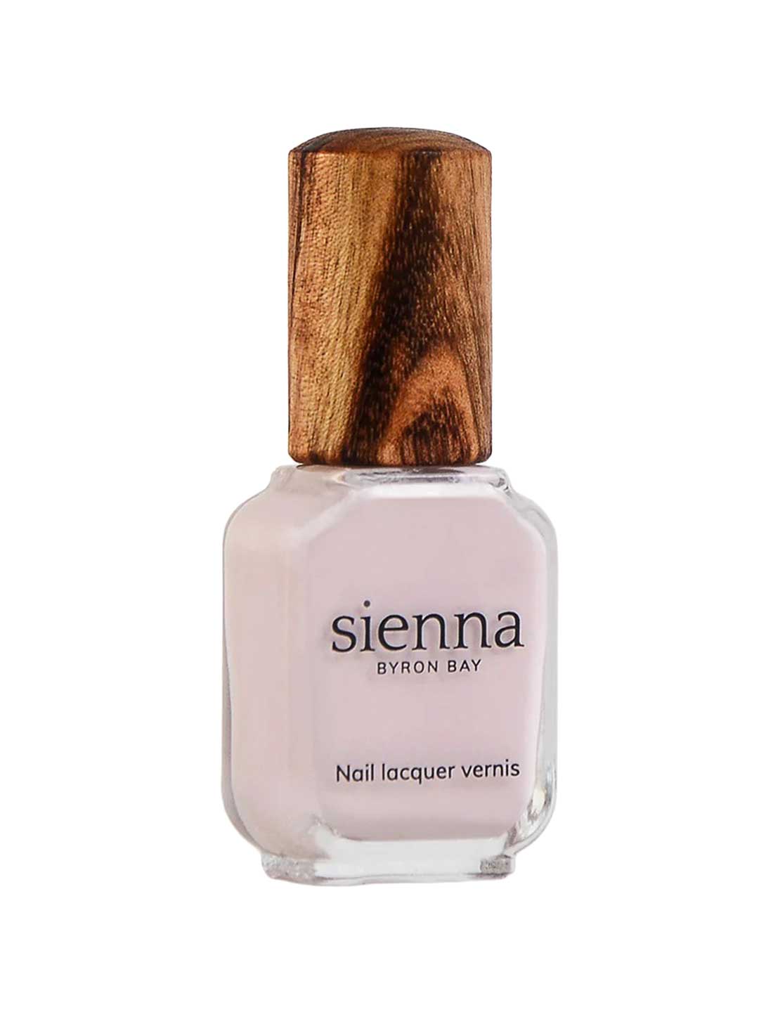 A bottle of Sienna Byron Bay Enlightened nail lacquer with pale pink polish, a clear rectangular glass base, and a textured wooden cap. The label reads “Sienna Byron Bay Nail lacquer vernis.”.