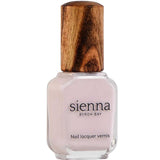 A bottle of Sienna Byron Bay Enlightened nail lacquer with pale pink polish, a clear rectangular glass base, and a textured wooden cap. The label reads “Sienna Byron Bay Nail lacquer vernis.”.