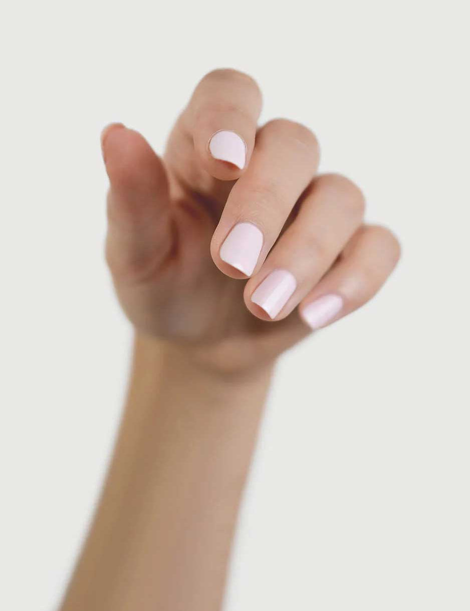 A hand with light skin tone and neatly manicured nails painted in Sienna Byron Bay Enlightened, a pale pink shade by Sienna Byron Bay, is shown with fingers slightly curled against a plain light background.