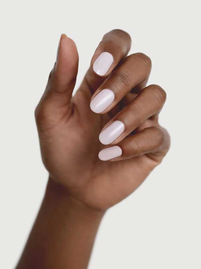 A hand with medium brown skin is shown against a light background, displaying neatly manicured nails painted in Sienna Byron Bay Enlightened, a glossy pale pink or white shade by Sienna Byron Bay.