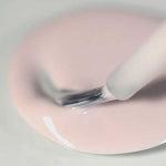 A close-up of a brush dipping into a pool of Sienna Byron Bay Enlightened, a light pink nail polish by Sienna Byron Bay, on a smooth surface.