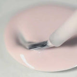 A close-up of a brush dipping into a pool of Sienna Byron Bay Enlightened, a light pink nail polish by Sienna Byron Bay, on a smooth surface.