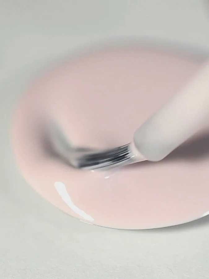 A close-up of a brush dipping into a pool of Sienna Byron Bay Enlightened, a light pink nail polish by Sienna Byron Bay, on a smooth surface.