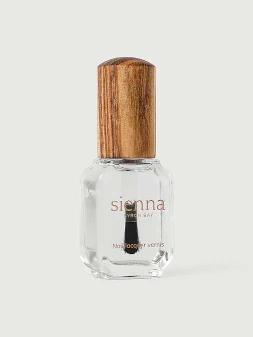 A clear glass bottle of Sienna Byron Bay Glossy Top Coat with a wooden cap, containing transparent polish, stands upright on a light gray background.