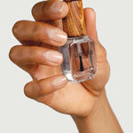A hand with well-groomed nails holds a clear bottle of Sienna Byron Bay Glossy Top Coat, featuring a wooden cap, against a light background.
