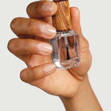 A hand with well-groomed nails holds a clear bottle of Sienna Byron Bay Glossy Top Coat, featuring a wooden cap, against a light background.