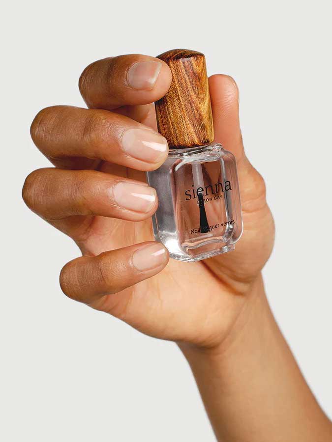 A hand with well-groomed nails holds a clear bottle of Sienna Byron Bay Glossy Top Coat, featuring a wooden cap, against a light background.