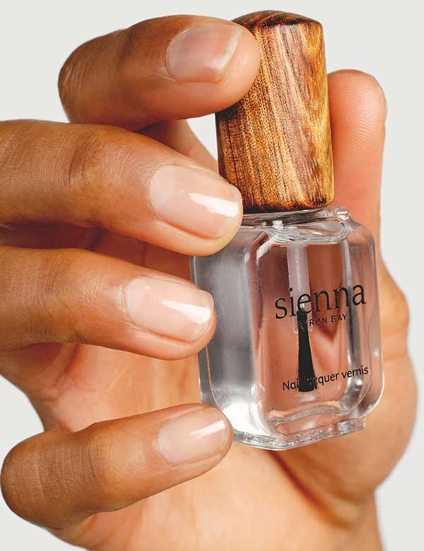 A hand holds a clear bottle of Sienna Byron Bay Glossy Top Coat with a wooden cap, displaying natural, clean nails.