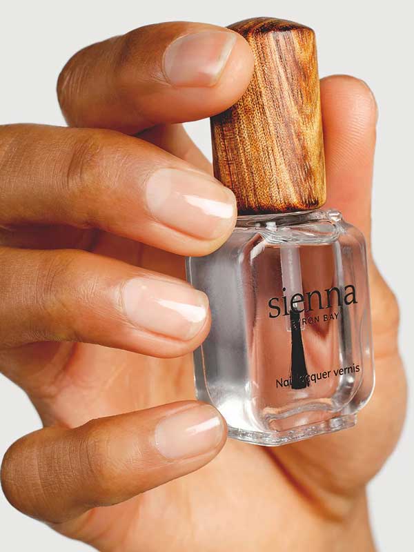 A hand holds a clear bottle of Sienna Byron Bay Glossy Top Coat with a wooden cap, displaying natural, clean nails.