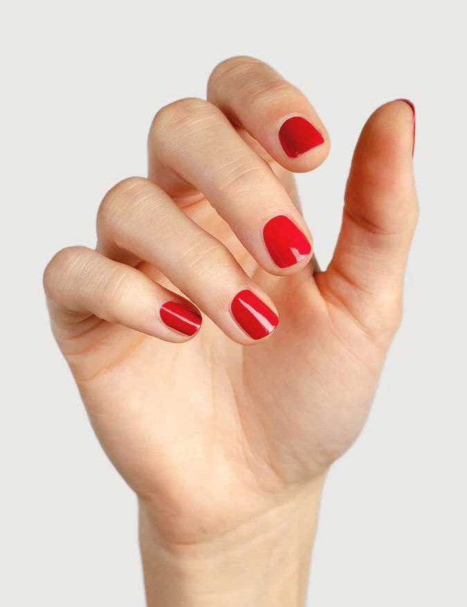 A hand with short, neatly filed nails painted in Sienna Byron Bay’s Heart, a glossy red polish, against a plain light background.