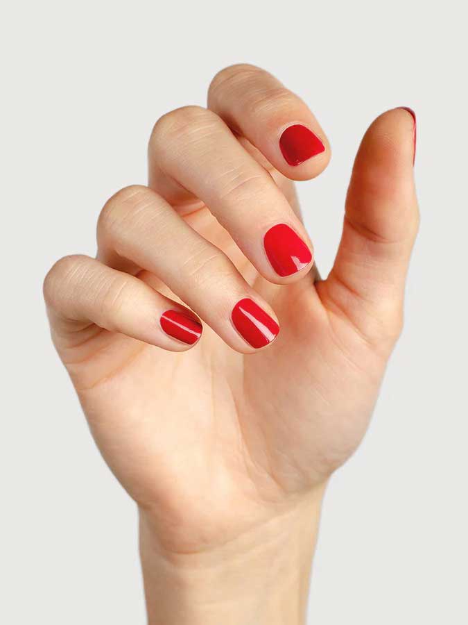 A hand with short, neatly filed nails painted in Sienna Byron Bay’s Heart, a glossy red polish, against a plain light background.