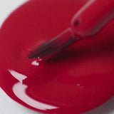 A close-up of a Sienna Byron Bay Heart nail polish brush resting on a puddle of glossy red polish from Sienna Byron Bay on a white surface.
