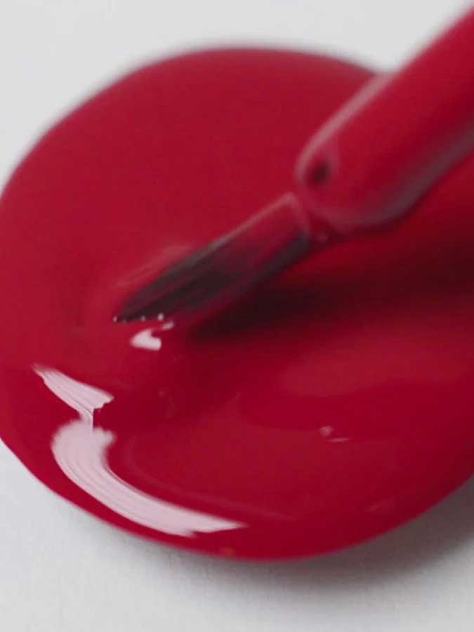 A close-up of a Sienna Byron Bay Heart nail polish brush resting on a puddle of glossy red polish from Sienna Byron Bay on a white surface.