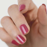 A close-up of a hand with short nails painted in Sienna Byron Bay Heartspace pink polish by Sienna Byron Bay, set against a plain, light background.