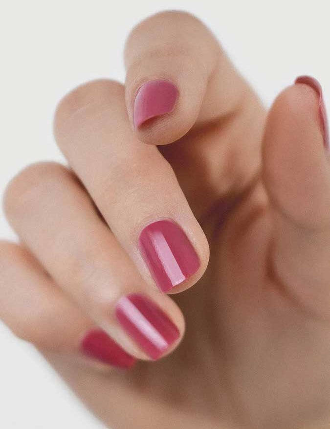A close-up of a hand with short nails painted in Sienna Byron Bay Heartspace pink polish by Sienna Byron Bay, set against a plain, light background.
