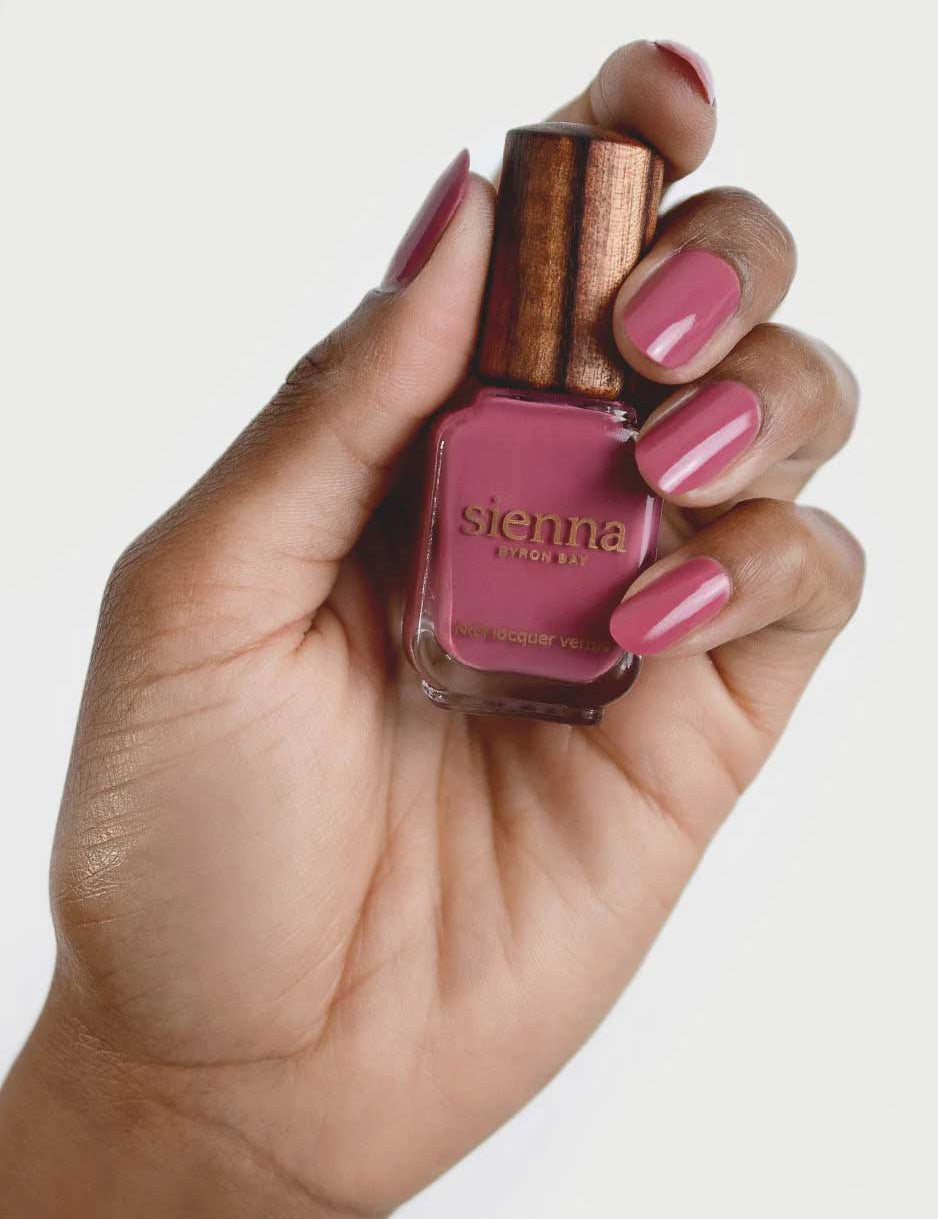 A hand with pink painted nails holds a bottle of Sienna Byron Bay Heartspace nail polish with a wooden cap, matching the nail color, against a plain white background.