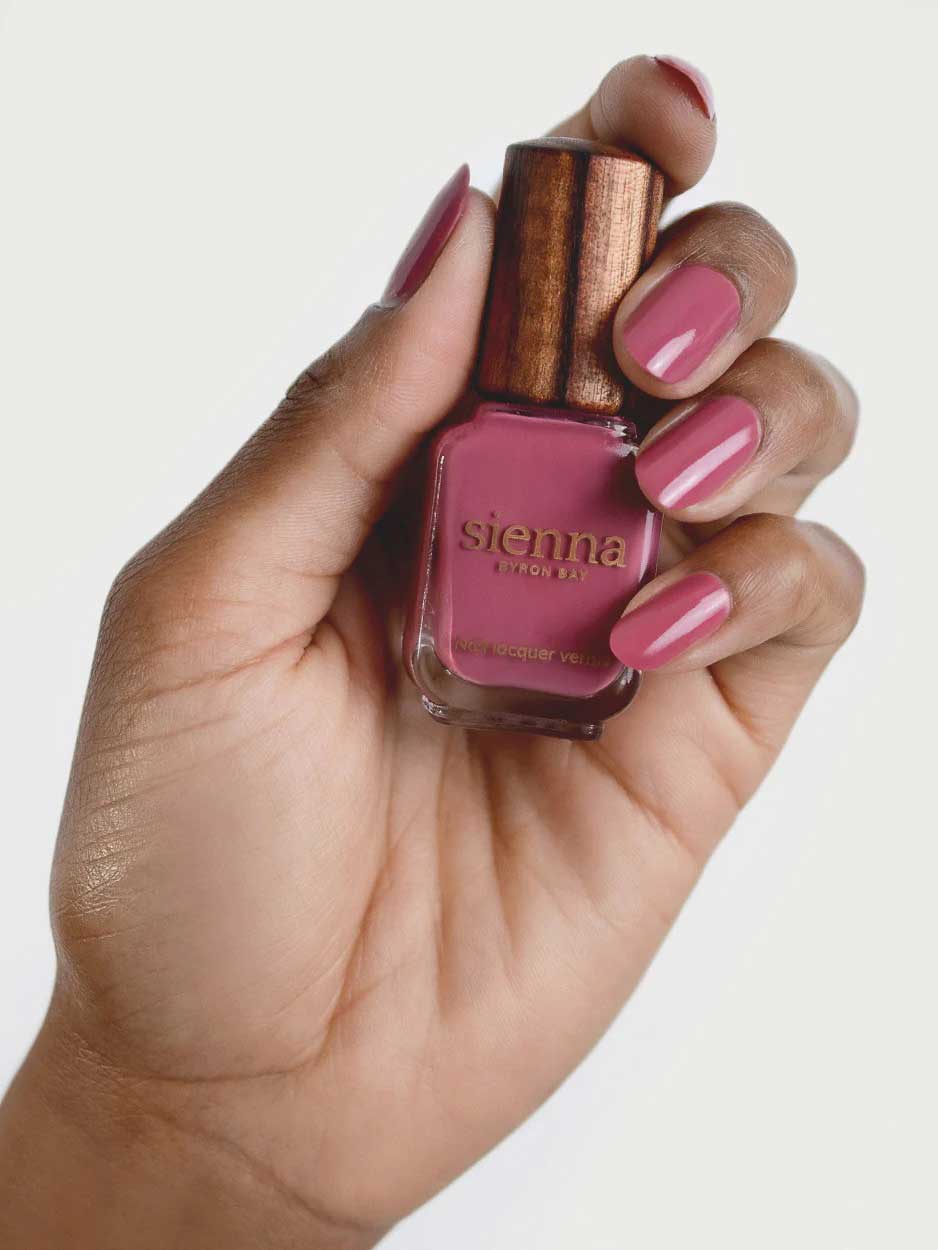 A hand with pink painted nails holds a bottle of Sienna Byron Bay Heartspace nail polish with a wooden cap, matching the nail color, against a plain white background.