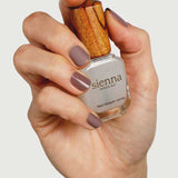 A hand with neatly manicured nails painted in matte mauve holds a bottle of Sienna Byron Bay Matte Top Coat with a wooden cap against a plain white background.