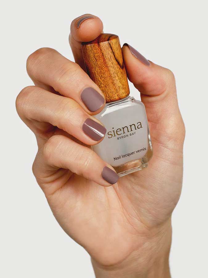 A hand with neatly manicured nails painted in matte mauve holds a bottle of Sienna Byron Bay Matte Top Coat with a wooden cap against a plain white background.