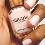 A hand with neatly manicured nails painted in pale pink holds a bottle of Sienna Byron Bay Melody nail lacquer, featuring the brand's signature wooden-textured cap.