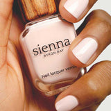 A hand with neatly manicured nails painted in pale pink holds a bottle of Sienna Byron Bay Melody nail lacquer, featuring the brand's signature wooden-textured cap.
