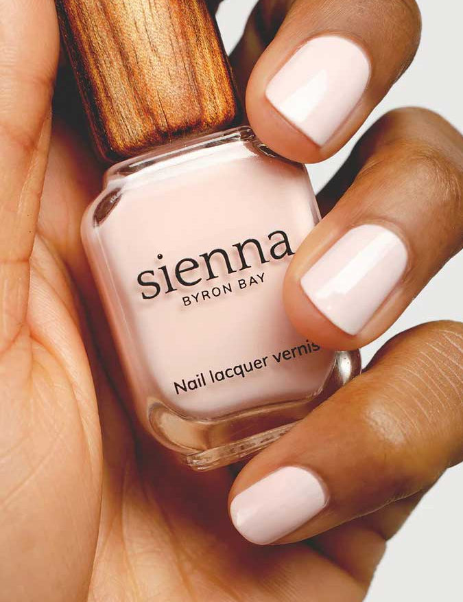 A hand with neatly manicured nails painted in pale pink holds a bottle of Sienna Byron Bay Melody nail lacquer, featuring the brand's signature wooden-textured cap.