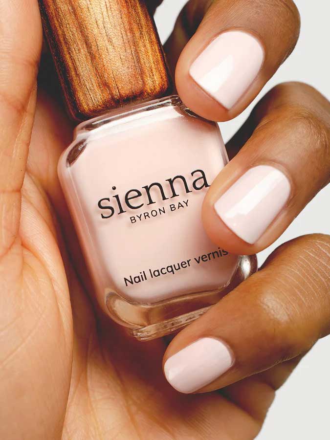 A hand with neatly manicured nails painted in pale pink holds a bottle of Sienna Byron Bay Melody nail lacquer, featuring the brand's signature wooden-textured cap.