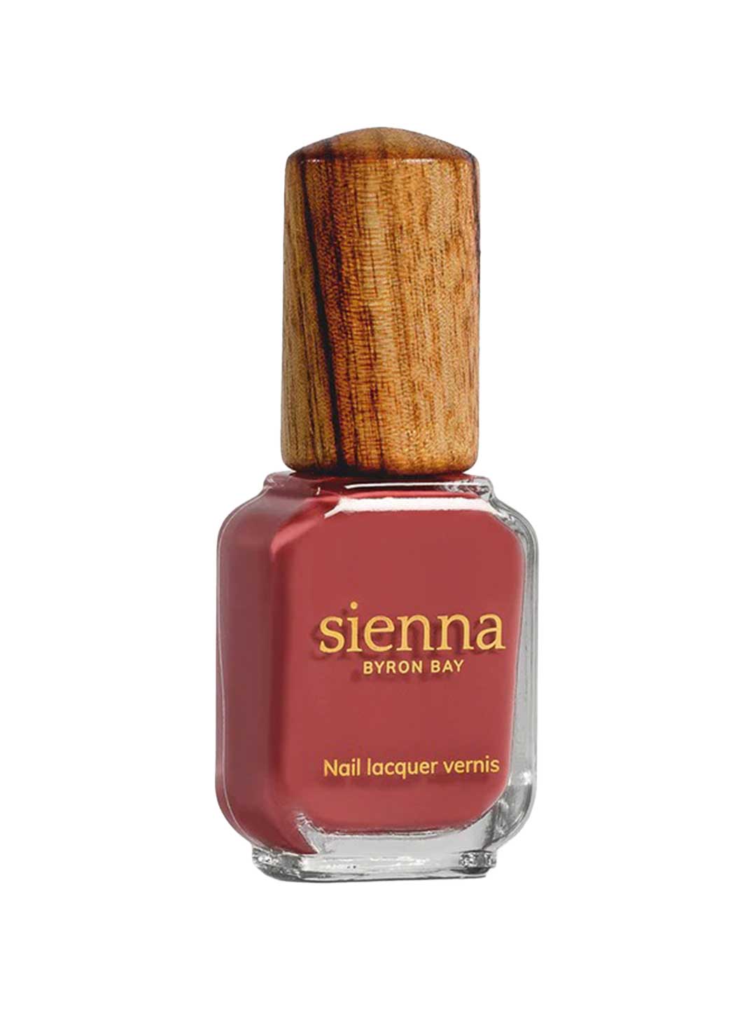 A bottle of Sienna Byron Bay Muse nail lacquer, a reddish-pink shade, features a unique wooden cap and gold text on the glass.