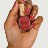 A hand with neatly manicured nails in a deep rose shade holds a Sienna Byron Bay Muse nail polish bottle with a wooden cap, set against a plain light background.