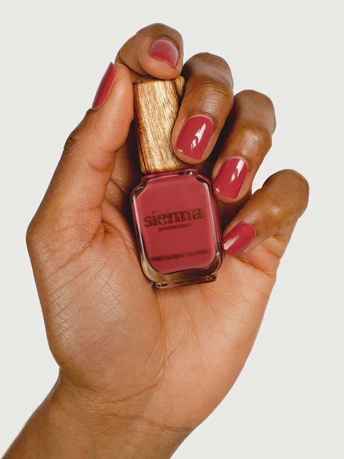 A hand with neatly manicured nails in a deep rose shade holds a Sienna Byron Bay Muse nail polish bottle with a wooden cap, set against a plain light background.