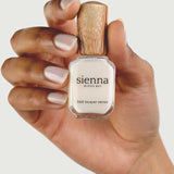 A hand with neatly manicured nails holds a bottle of Sienna Byron Bay - Hope nail lacquer with a wooden cap against a light background.