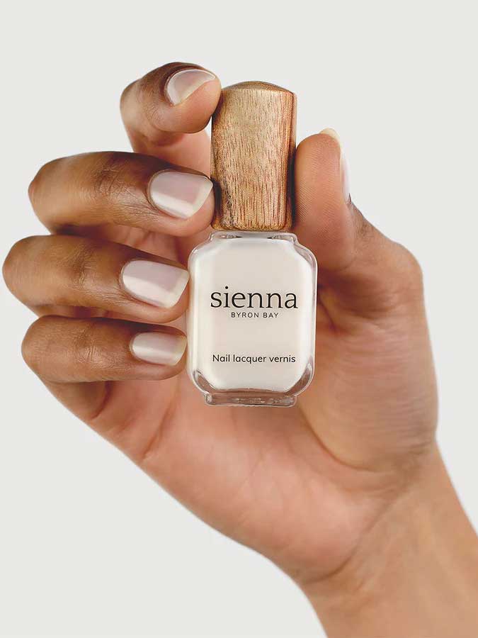 A hand with neatly manicured nails holds a bottle of Sienna Byron Bay - Hope nail lacquer with a wooden cap against a light background.