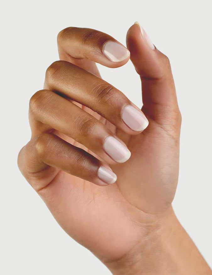 A hand with medium brown skin tone is shown against a plain background, fingers slightly curled. The neatly manicured nails are painted with Sienna Byron Bay - Hope by Sienna Byron Bay, a pale, natural-looking polish.