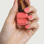 A hand holds a bottle of Sienna Byron Bay Laughter nail polish with a wooden cap, displaying neatly manicured nails painted in the same cheerful coral pink by Sienna Byron Bay.