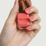 A hand holds a bottle of Sienna Byron Bay Laughter nail polish with a wooden cap, displaying neatly manicured nails painted in the same cheerful coral pink by Sienna Byron Bay.