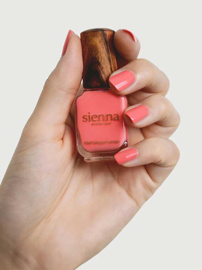 A hand holds a bottle of Sienna Byron Bay Laughter nail polish with a wooden cap, displaying neatly manicured nails painted in the same cheerful coral pink by Sienna Byron Bay.
