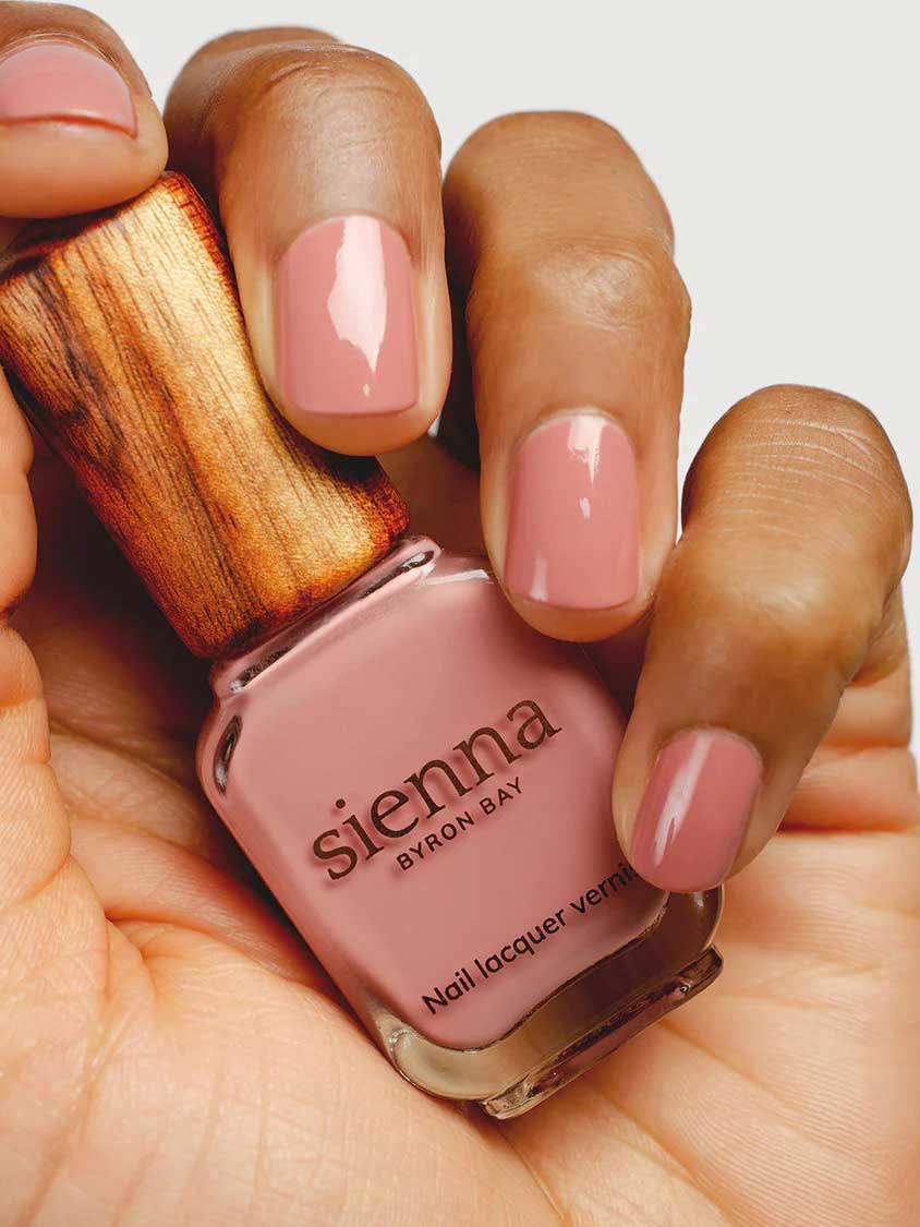 A hand with neatly manicured nails painted in a soft pink shade holds a bottle of Sienna Byron Bay Nail Polish - Stone with a wooden cap.