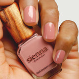 A hand with neatly manicured nails painted in a soft pink shade holds a bottle of Sienna Byron Bay Nail Polish - Stone with a wooden cap.