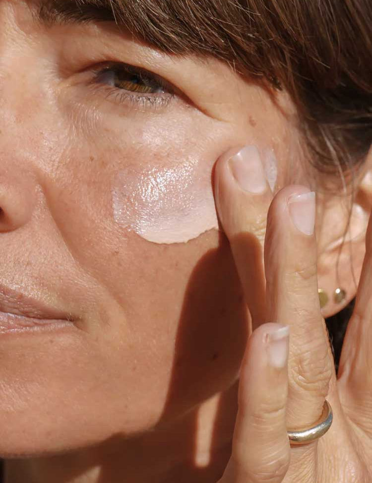 Close-up of a person with light skin and brown hair applying Mukti Daily Moisturiser with Sunscreen Solar Veil SPF30 100ml under their eye, gently blending it in with a finger. The focus is on the eye and cheek area; a silver ring is visible.