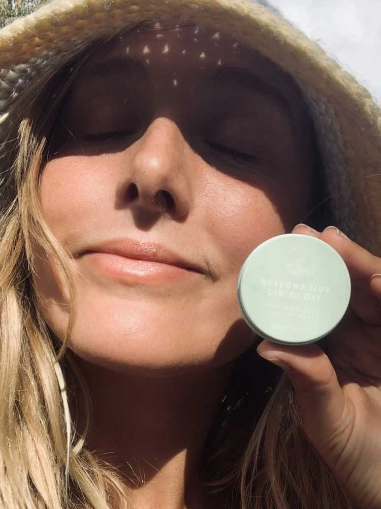 A woman wearing a straw hat smiles with her eyes closed in sunlight, holding a small round container labeled SunButter Skincare Restorative Lip Gloss 15ml near her face.