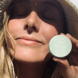 A woman wearing a straw hat smiles with her eyes closed in sunlight, holding a small round container labeled SunButter Skincare Restorative Lip Gloss 15ml near her face.