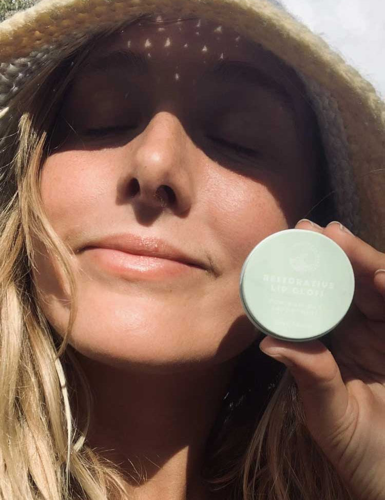 A woman wearing a straw hat smiles with her eyes closed in sunlight, holding a small round container labeled SunButter Skincare Restorative Lip Gloss 15ml near her face.