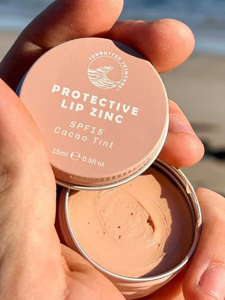 A hand holds an open tin of SunButter Skincare SPF15 Protective Lip Zinc – Cacao Tint 15ml, displaying the creamy zinc oxide formula inside against a beach background.