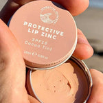A hand holds an open tin of SunButter Skincare SPF15 Protective Lip Zinc – Cacao Tint 15ml, displaying the creamy zinc oxide formula inside against a beach background.