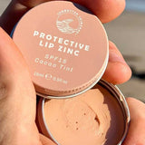 A hand holds an open tin of SunButter Skincare SPF15 Protective Lip Zinc – Cacao Tint 15ml, displaying the creamy zinc oxide formula inside against a beach background.