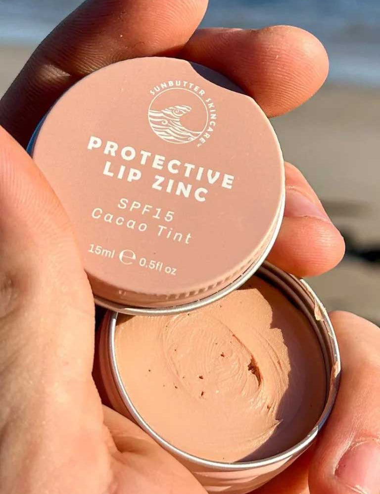 A hand holds an open tin of SunButter Skincare SPF15 Protective Lip Zinc, Cacao Tint 15ml, displaying the creamy zinc oxide formula inside against a beach background.