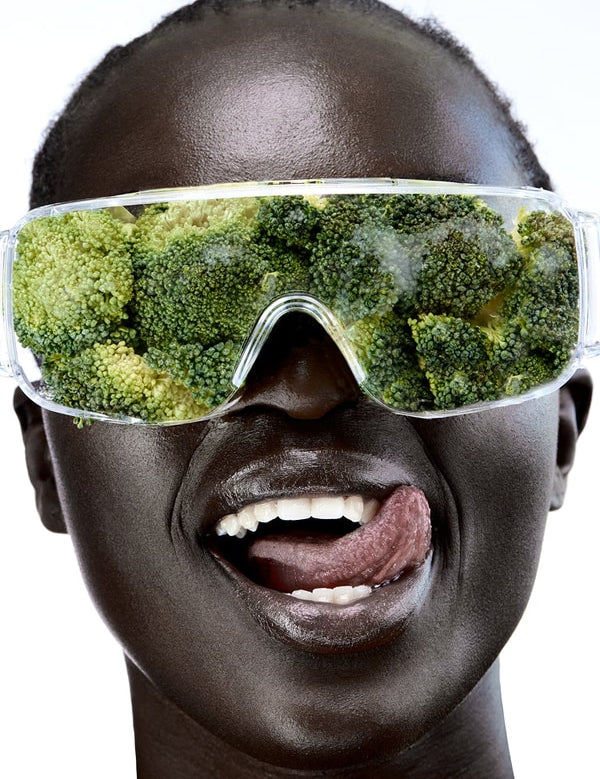 Close-up of a smiling person wearing clear wraparound goggles filled with broccoli florets, with their tongue touching the corner of their mouth.